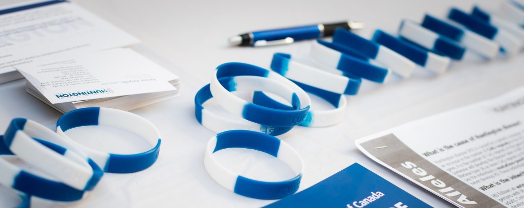 A display of promotional materials on a white tablecloth. The display includes a stack of business cards with a Huntington Society of Canada logo, a row of blue and white silicone awareness bracelets, a blue pen, and paper handouts.